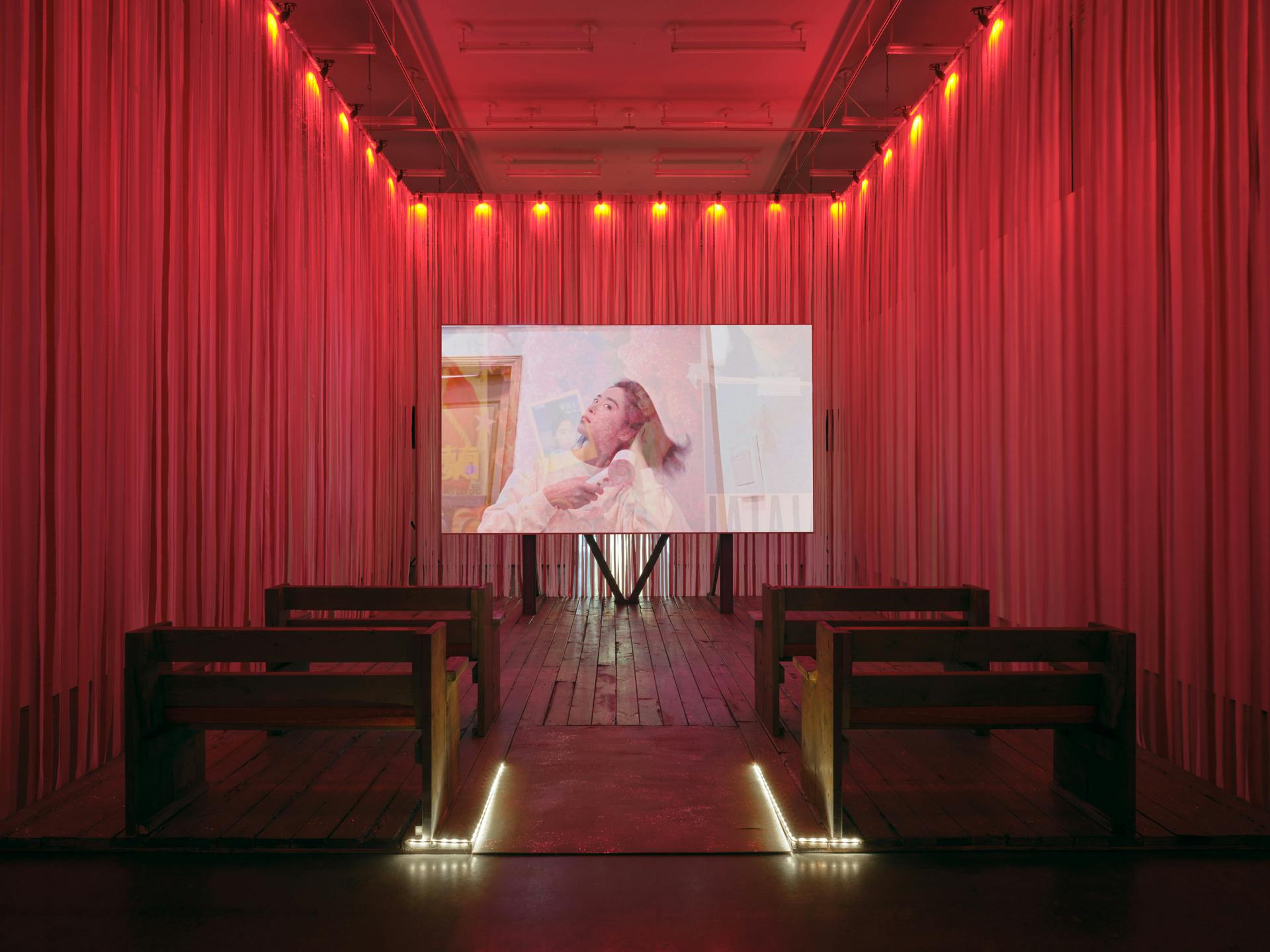 Two rows of benches face a screen in a room with pink and red ribbons walls. Onscreen, an image of a woman blowdrying her hair is overlain on a collage made of movie paraphernalia.