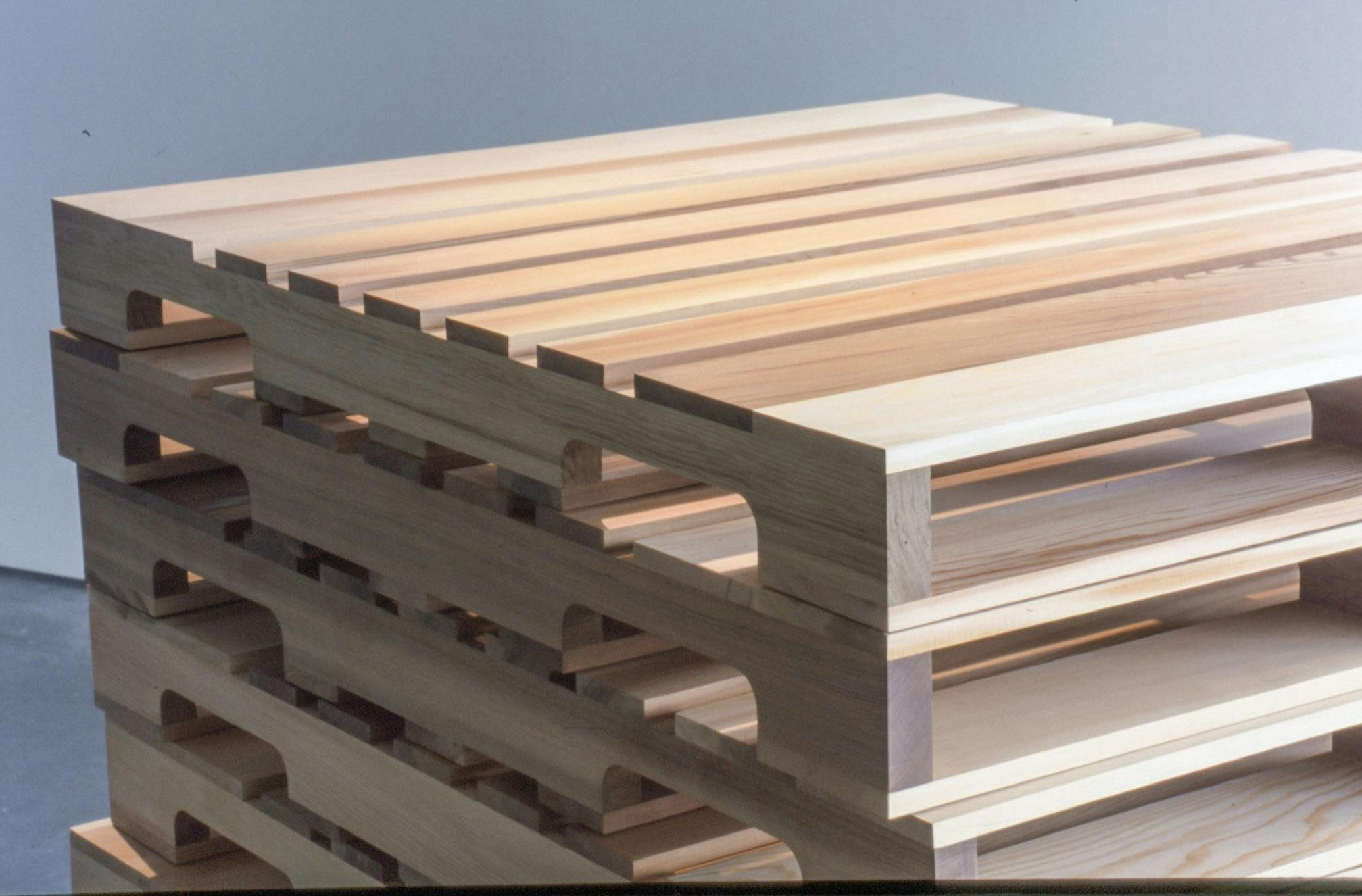 A stack of wood pieces, which look like large duckboards, is installed in a gallery space. They are identical in shape and size. This close-up photograph shows only the top five boards on this stack. 