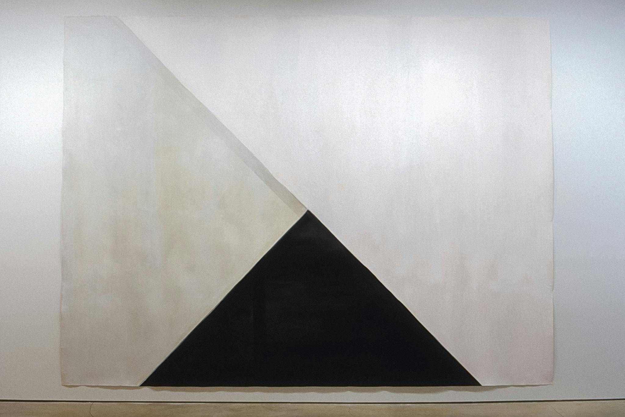 A large painting is installed on a gallery wall. It contains three geometric shapes: above the black triangle at the bottom, two white trapezoids are placed partially overlapping with each other. 