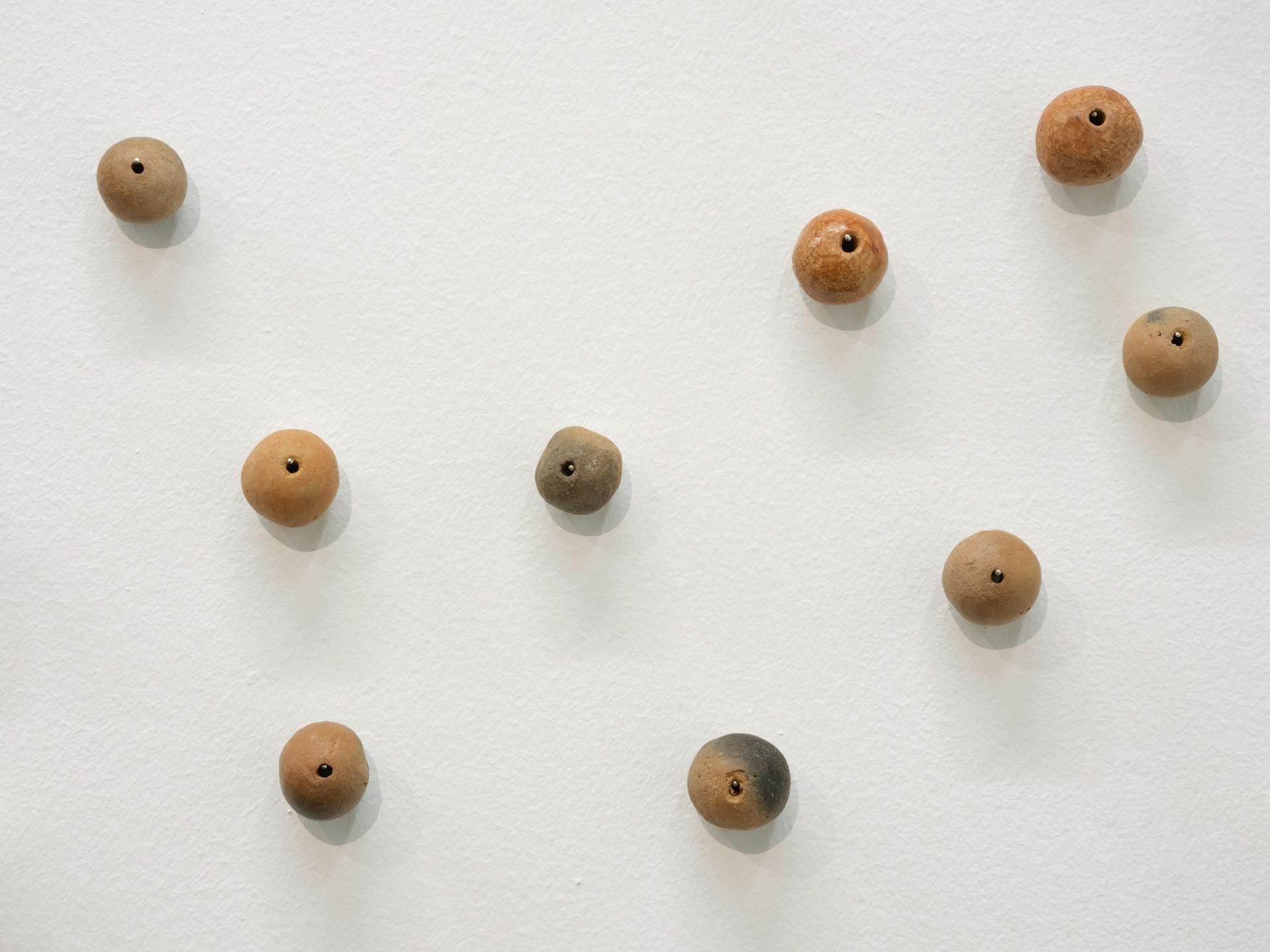 Detail of nine clay beads in orange and grey tones pinned to a white wall.