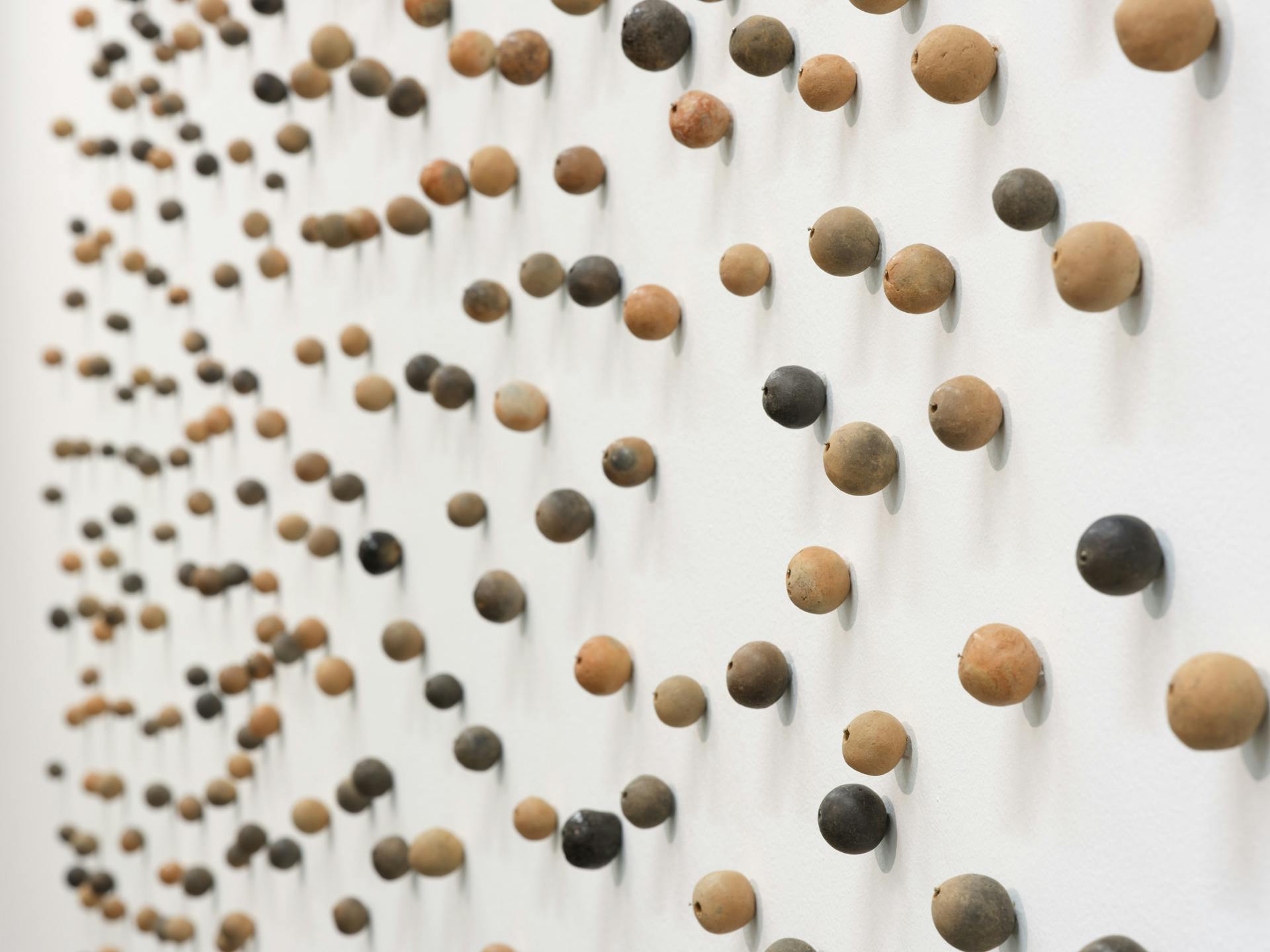 Hundreds of small clay beads of various colours are pinned to a white wall with varying distances between each bead.