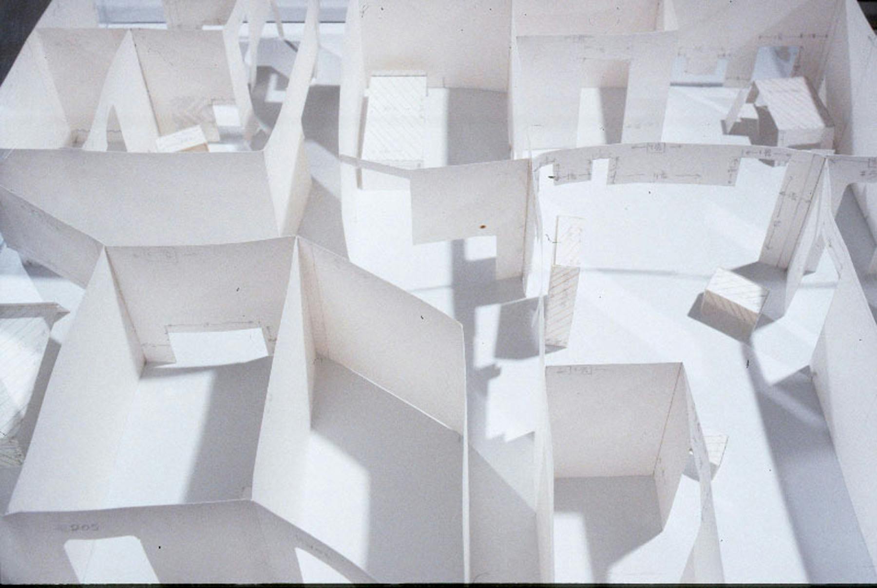 A close view of a paper sculpture. The sculpture is a maquette of a fictitious apartment with many rooms. Annotations and measurements are visible on some of the walls.