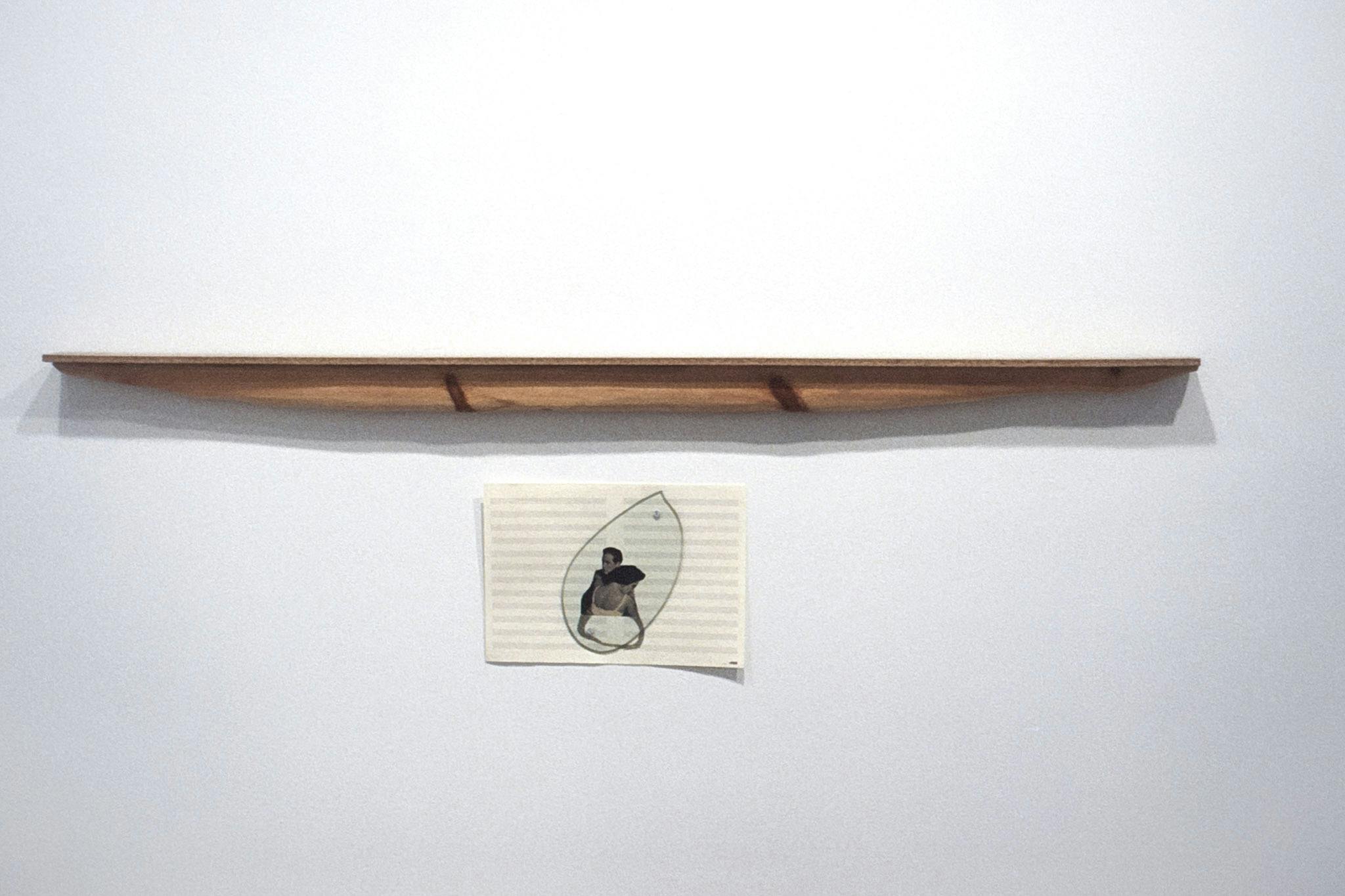 A wooden shelf-like sculpture hangs horizontally on a wall. A drawing of two figures embracing made on blank musical notation paper is pinned to the wall beneath it. 