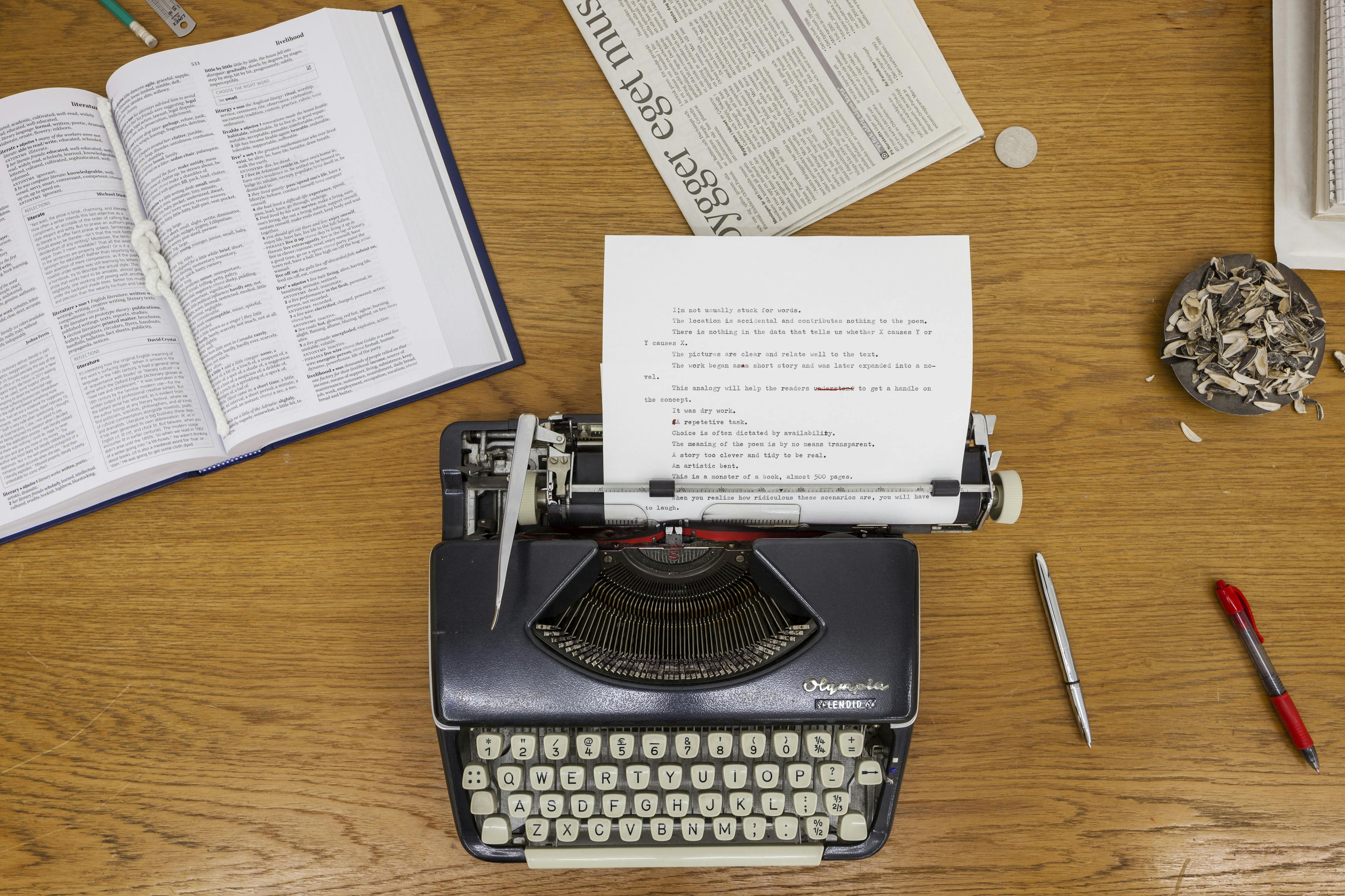 An overhead view of a black typewriter on a wooden desk surrounded by open books, papers, pens and a bowl of sunflower seed shells.