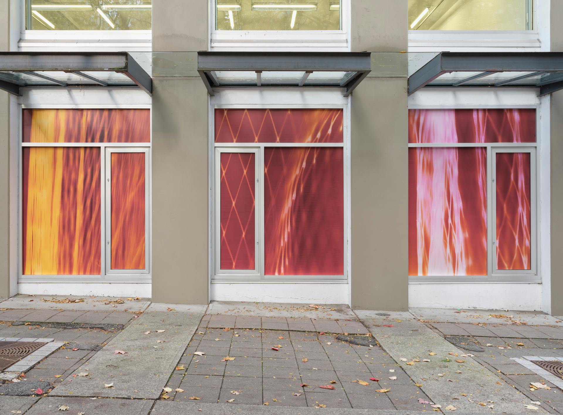 Three windows featuring three photograms depicting nylon mesh produce bags against neon photographic paper. From left to right the photograms colours' vary from orange to pink and red.