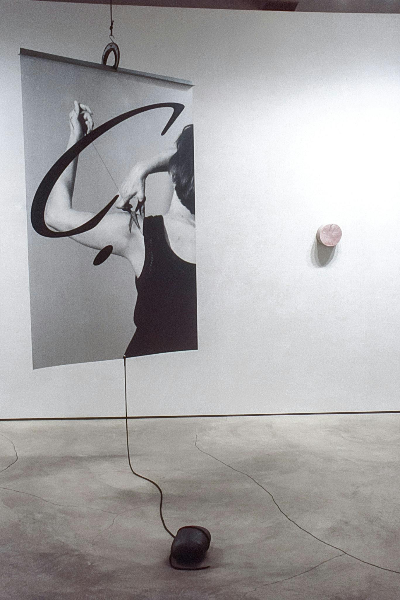 An artwork suspended in a gallery space. The work. is composed of a large photo that shows someone cutting a thread from their tank top. A rolled cable hangs from the bottom of the photo. 