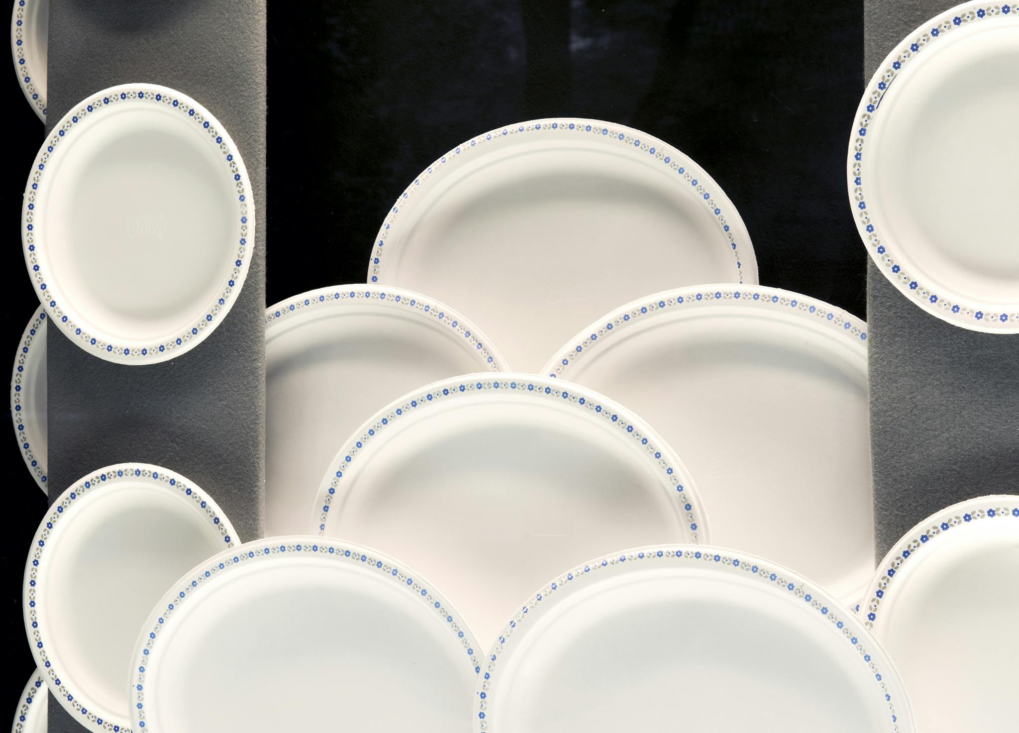 A close up view of an installation containing white paper plates edged with a blue floral pattern. The plates are placed upright over a black background and layered creating a scalloped pattern. 