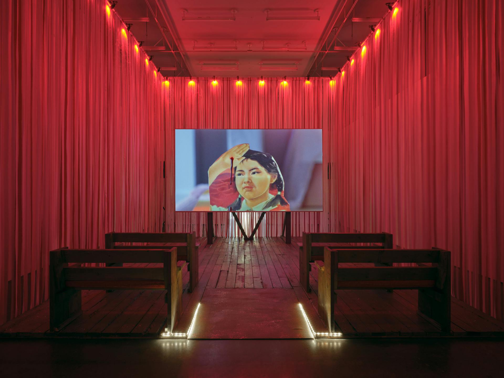 Two rows of benches face a screen in a room with pink and red ribbons walls. Onscreen, red liquid drips down the palm of a paper cut-out propaganda-style image of a young girl saluting. 