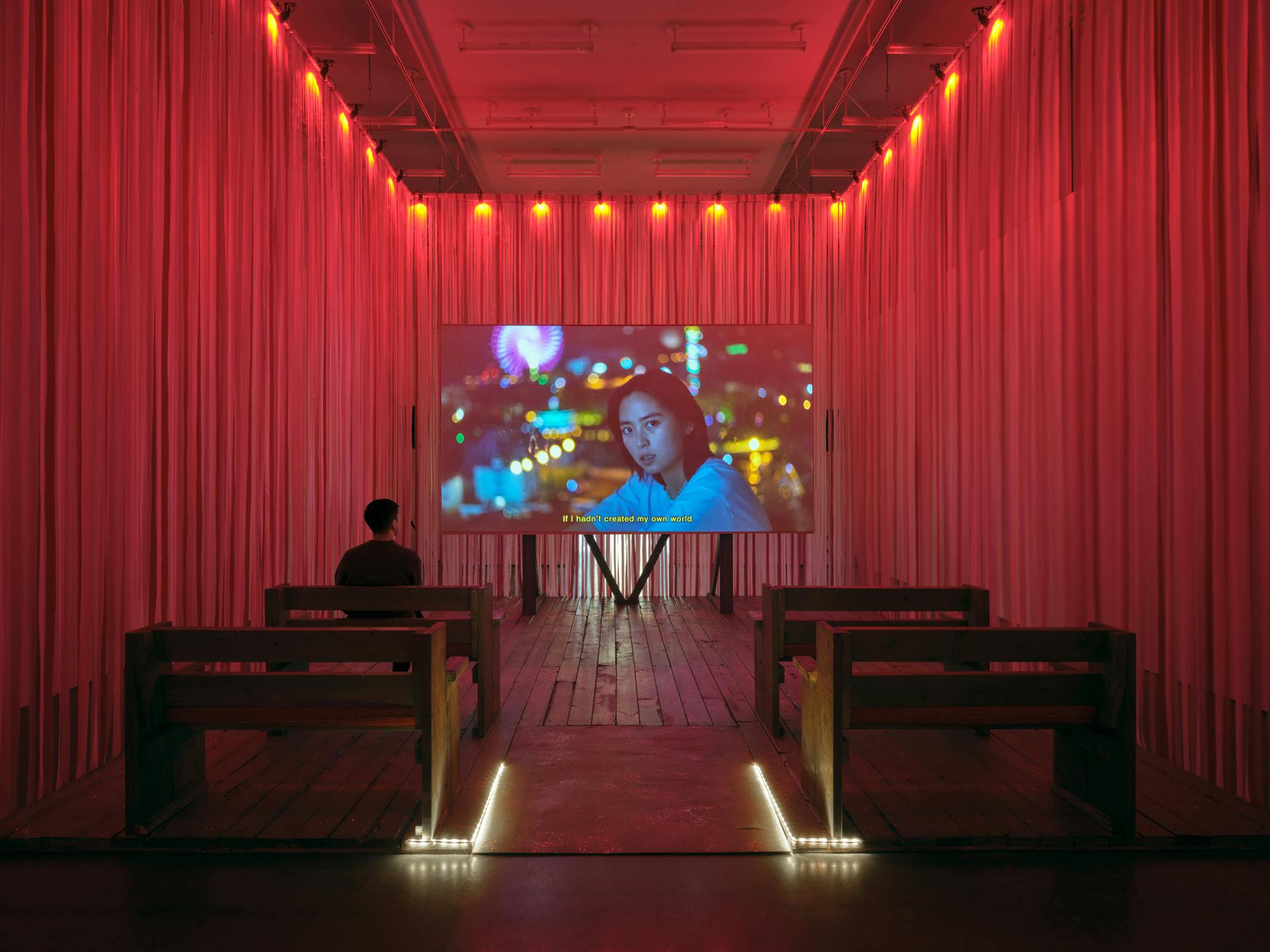 Two rows of benches face a screen in a room with pink and red ribbons walls. Onscreen, a woman faces the viewer with the text caption If I hadn’t created my own world at the bottom of the screen. 
