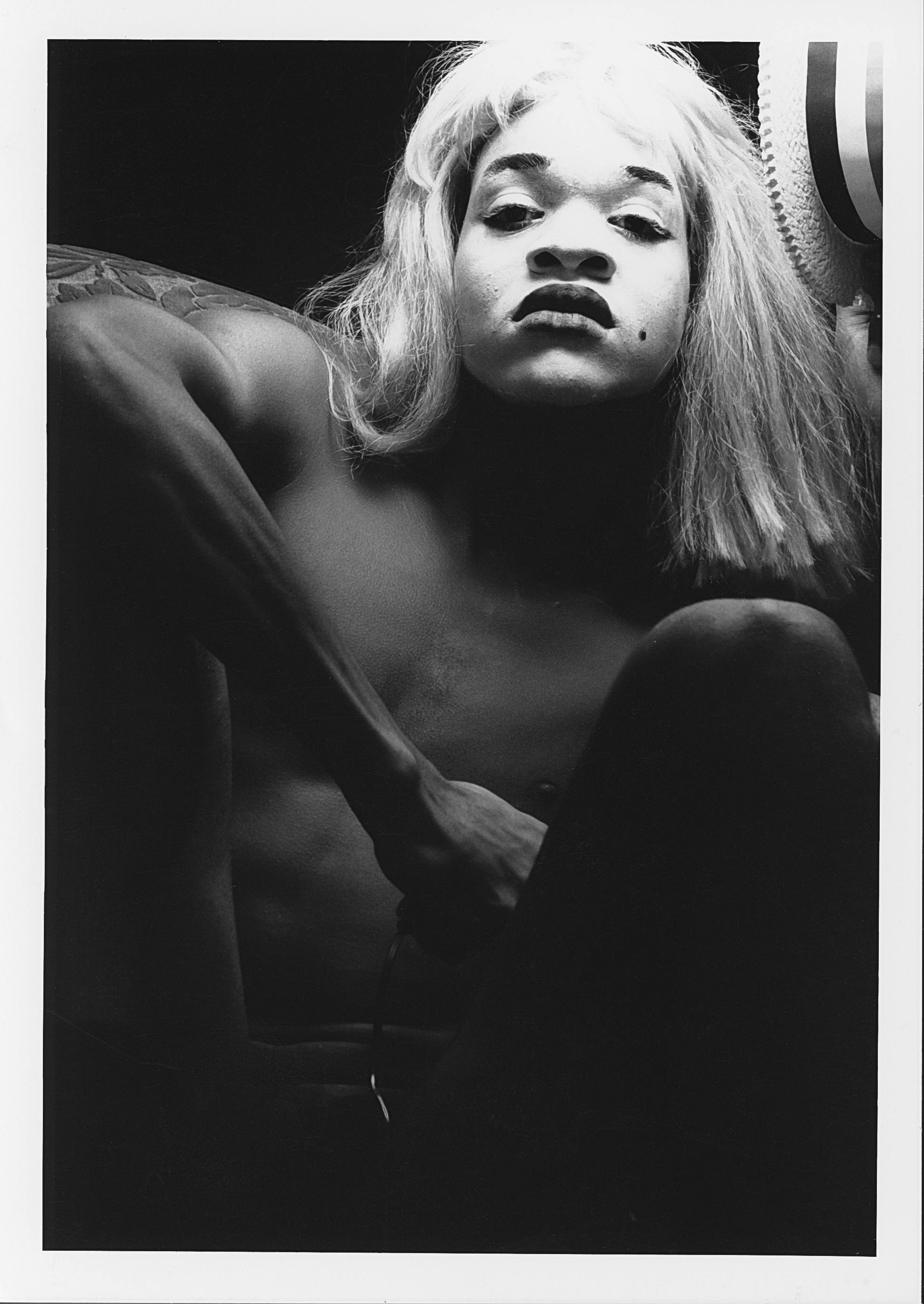 A starkly lit black and white portrait of a courching shirtless person holding a photo release trigger in their hand. They are wearing white face makeup and a wig. 