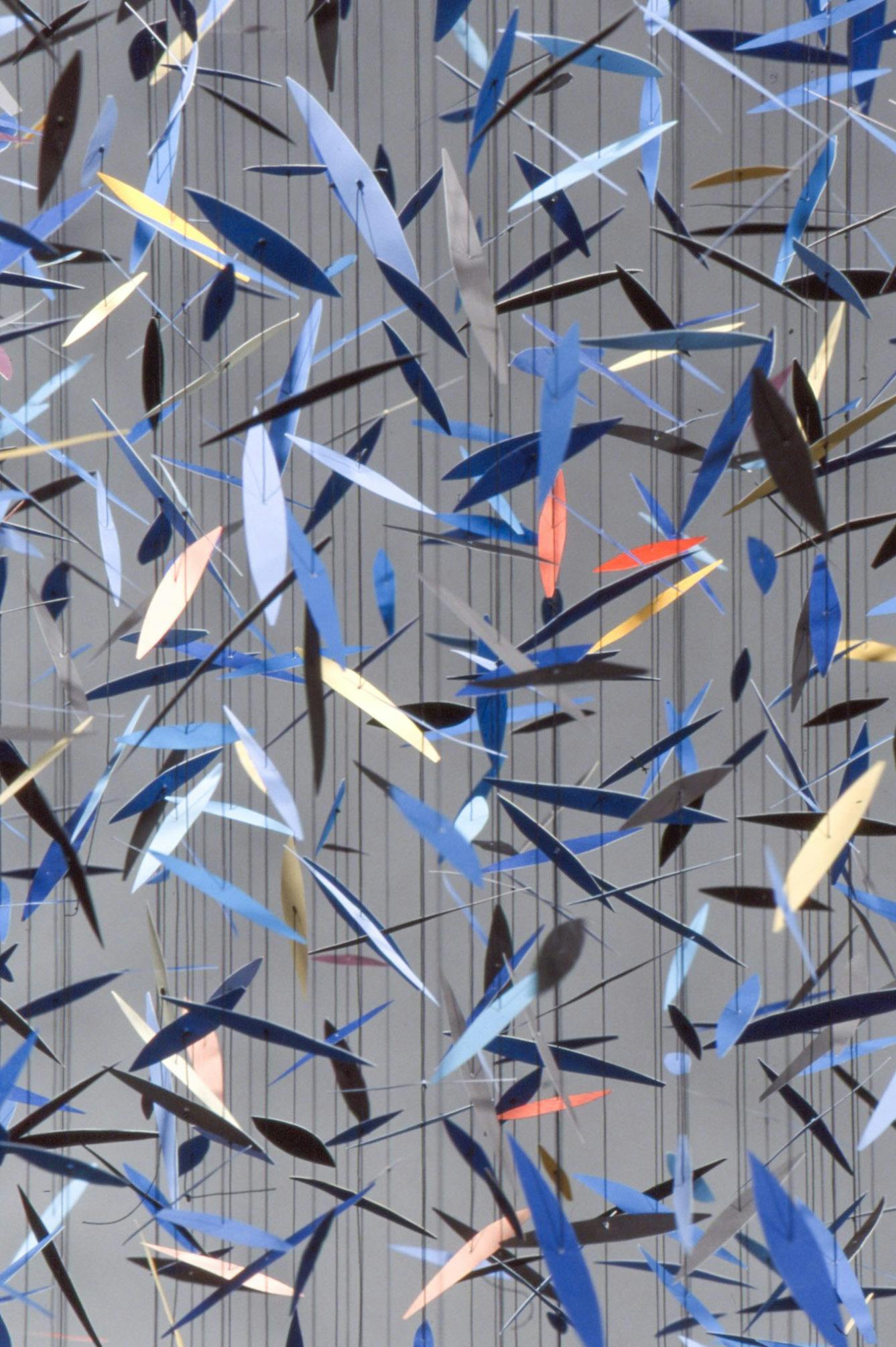 This is a close-up view of a sculptural installation by Pae White. A countless number of small leaf-shaped blue and orange papers are hanging from the ceiling through black threads. 