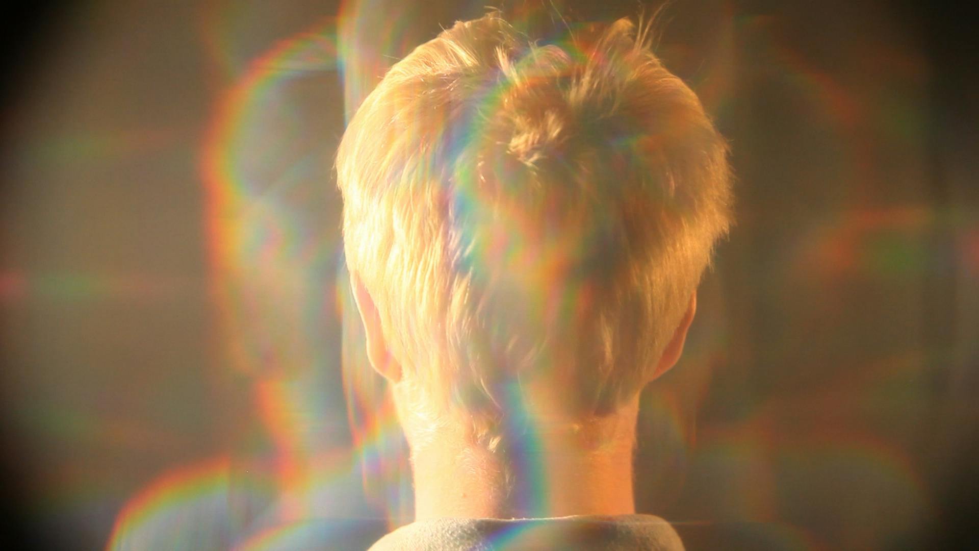 An image depicting the back of a person’s head, with short blonde hair. The person is cast in warm light with rainbows refracting throughout the image. 