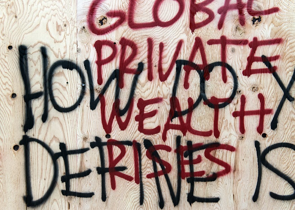 A close up image of overlapping spray-painted words in red and black on a wooden surface. One phrase reads, “GLOBAL PRIVATE WEALTH RISES,” the other is fragmented and reads “HOW DO Y DEFINE IS.” 