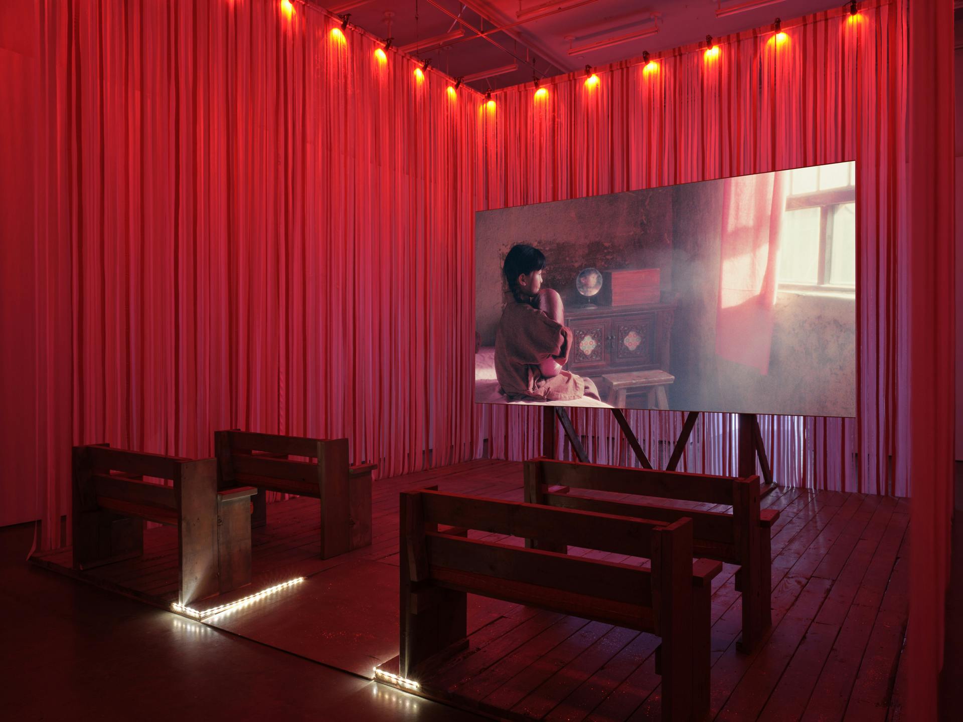 Diagonal view of four benches facing a large screen with pink and red ribbons forming a curtain around the perimeter. Onscreen, a child sits on a bed in a dark room, clutching a pink object and looking out a window.