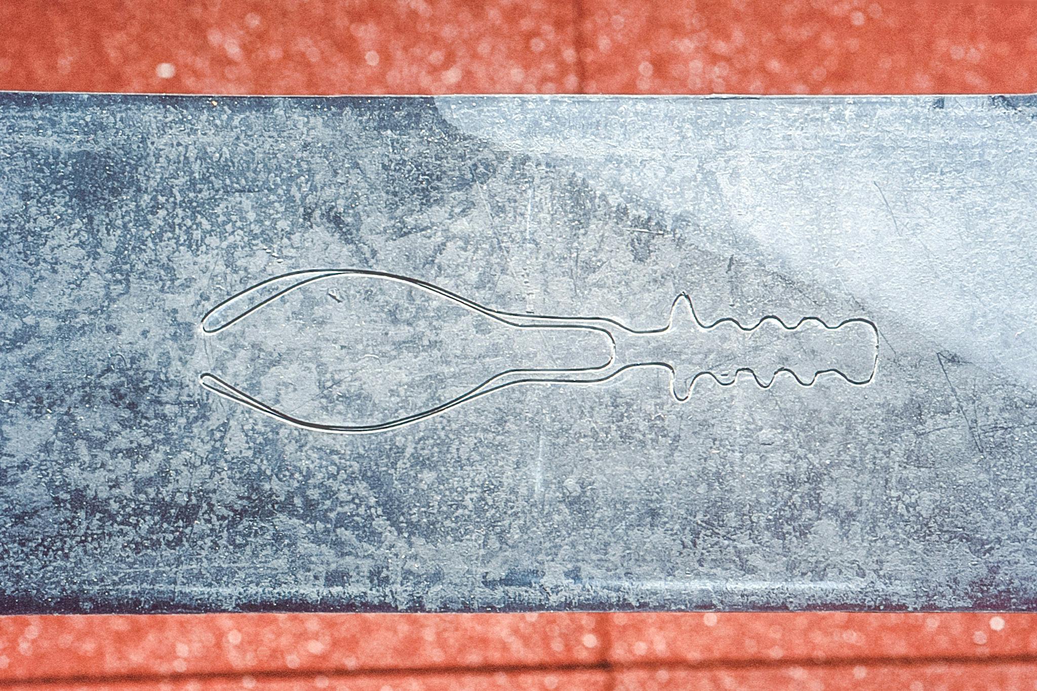 An incomplete shape of a children’s hand shovel is carved on the metal surface. The handle is fully visible, but the blade is half recognizable as if it is stuck in the sand.