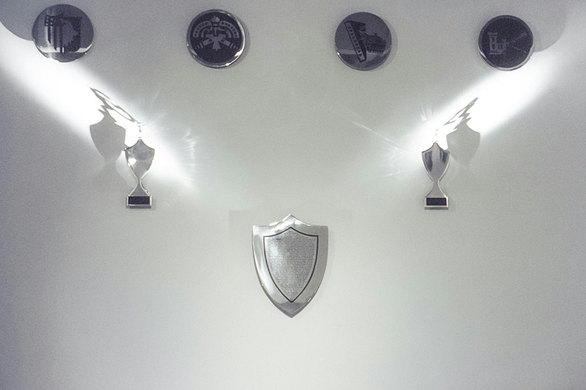 Several artworks on a white wall. At the top, there's a row of 4 metal circles with black crests. Below, there are 2 small trophy shapes . At the bottom center there is a shield shape with small text.