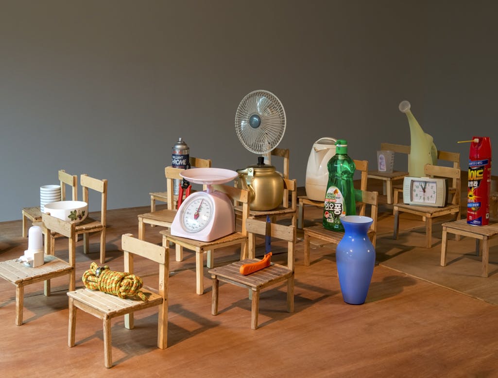Miniature chairs placed in rows are placed atop a wooden surface. Various objects such as a kitchen scale, fan, rope, dish soap bottle, clock, bug spray, kettle and watering can sit on the chairs.