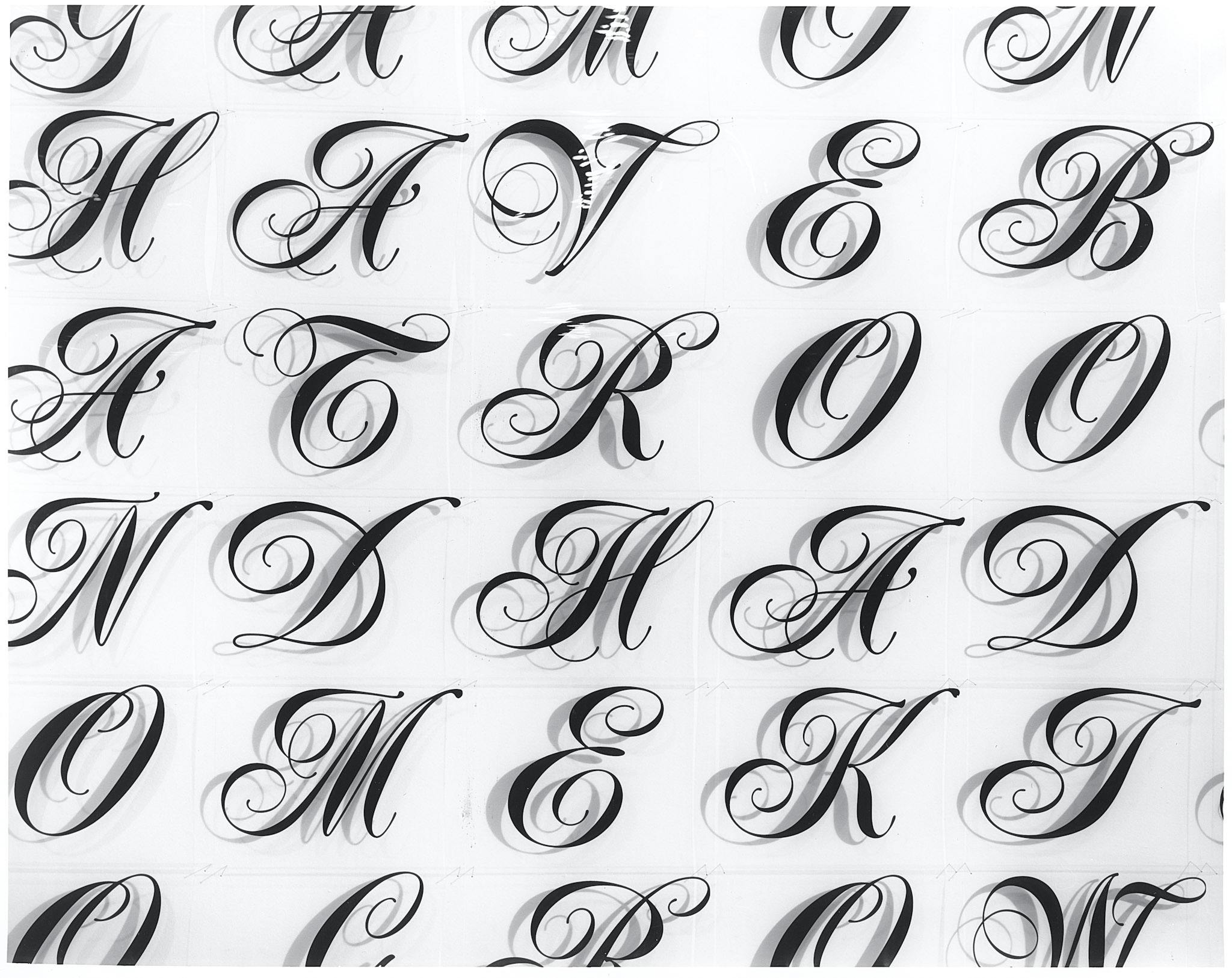 A closeup of several cursive capital letters. The letters are printed on clear plastic film and pasted on white walls, creating repeating shadows that replicate their curves and details.